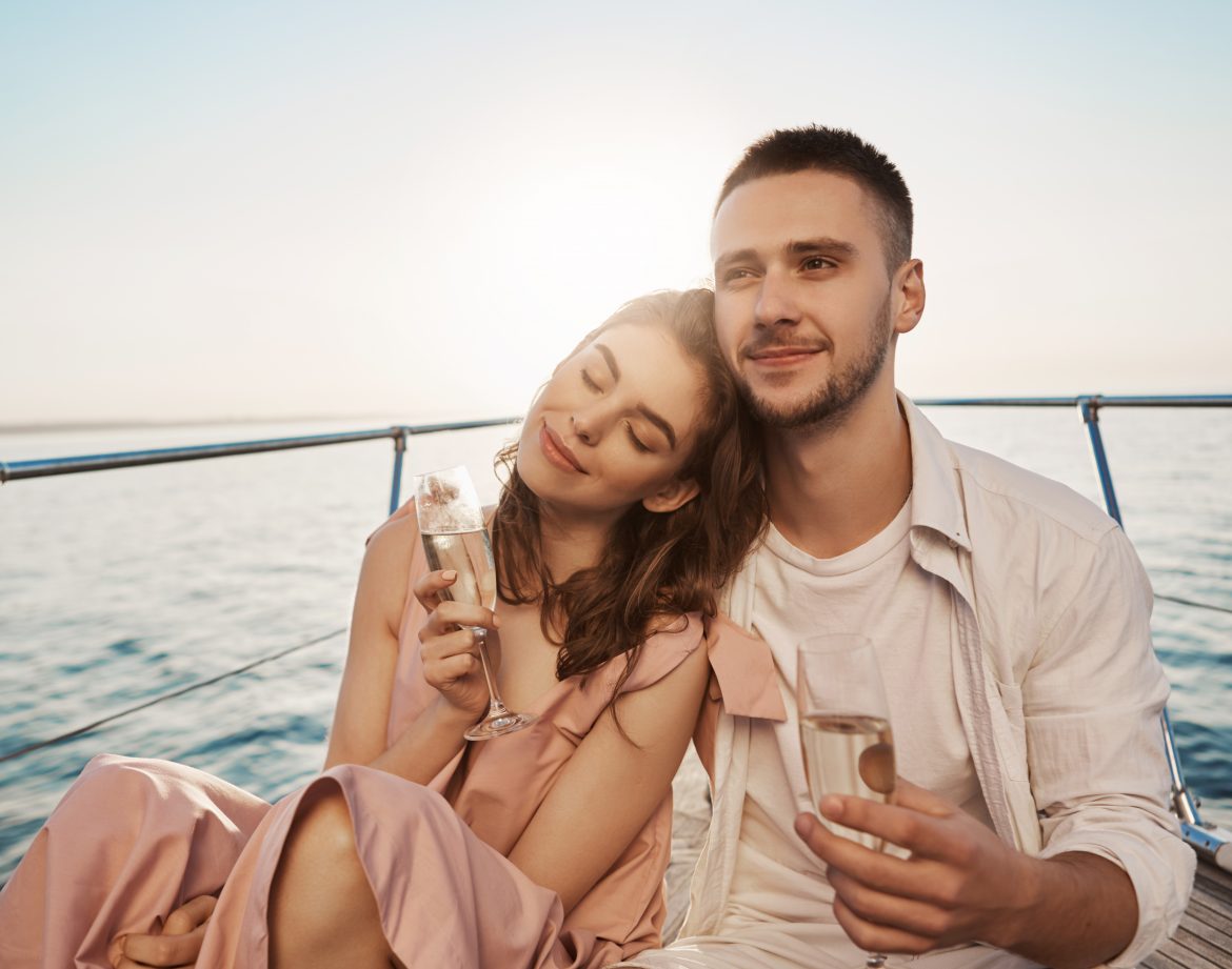Outdoor portrait of adult happy male and female in love, celebrating their engagement on yacht, holding glass of champagne and hugging. Boyfriend just told about things he likes in her. Emotions and vacation concept.