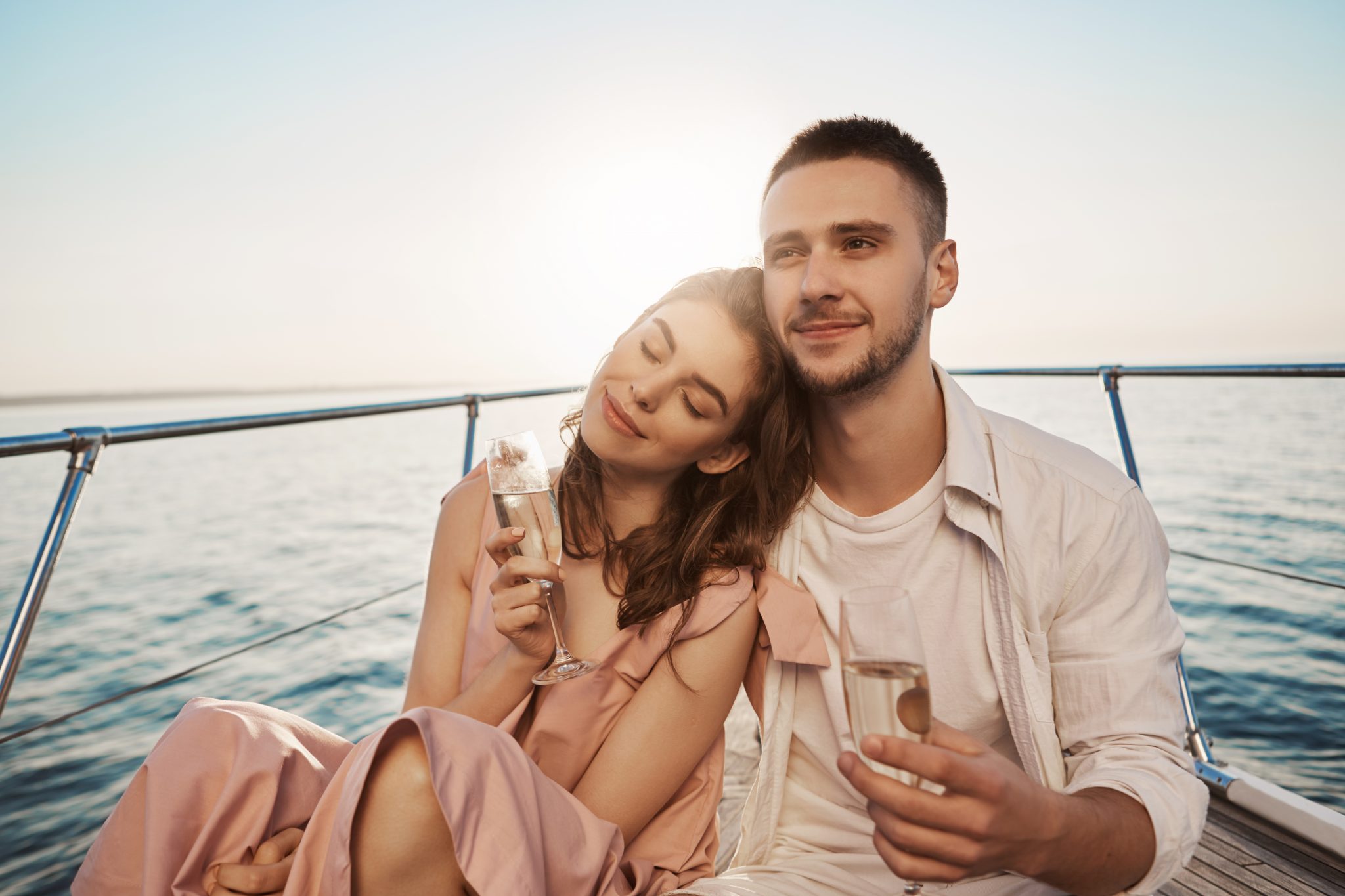 Outdoor portrait of adult happy male and female in love, celebrating their engagement on yacht, holding glass of champagne and hugging. Boyfriend just told about things he likes in her. Emotions and vacation concept.